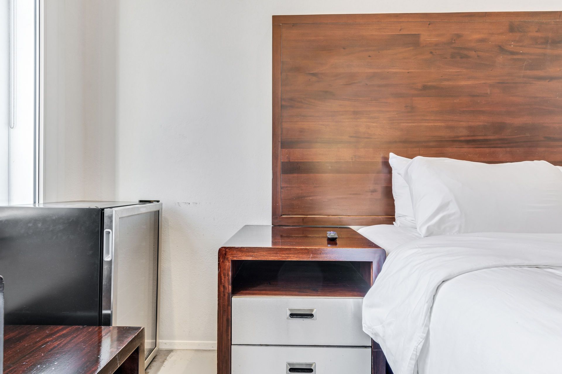Bedroom with a bed, nightstand, and small refrigerator. White bedding, brown headboard and furniture.