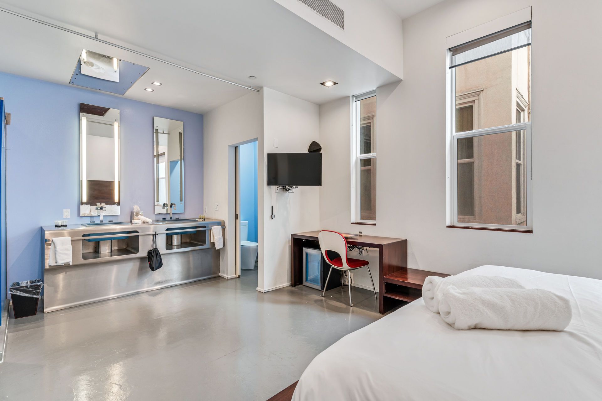 Modern hotel room with open bathroom, desk, bed, and natural light from large windows.