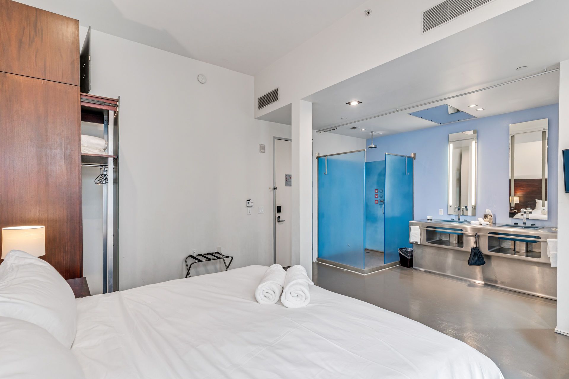 Modern hotel room with bed, open bathroom with blue shower, and two sinks.