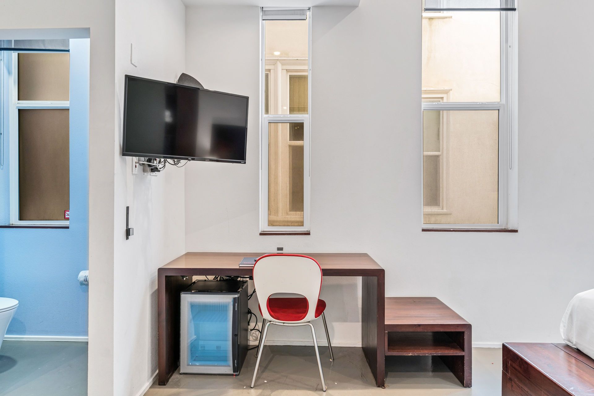 A hotel room with a desk, chair, TV, and mini-fridge against a white wall; bathroom on the left.