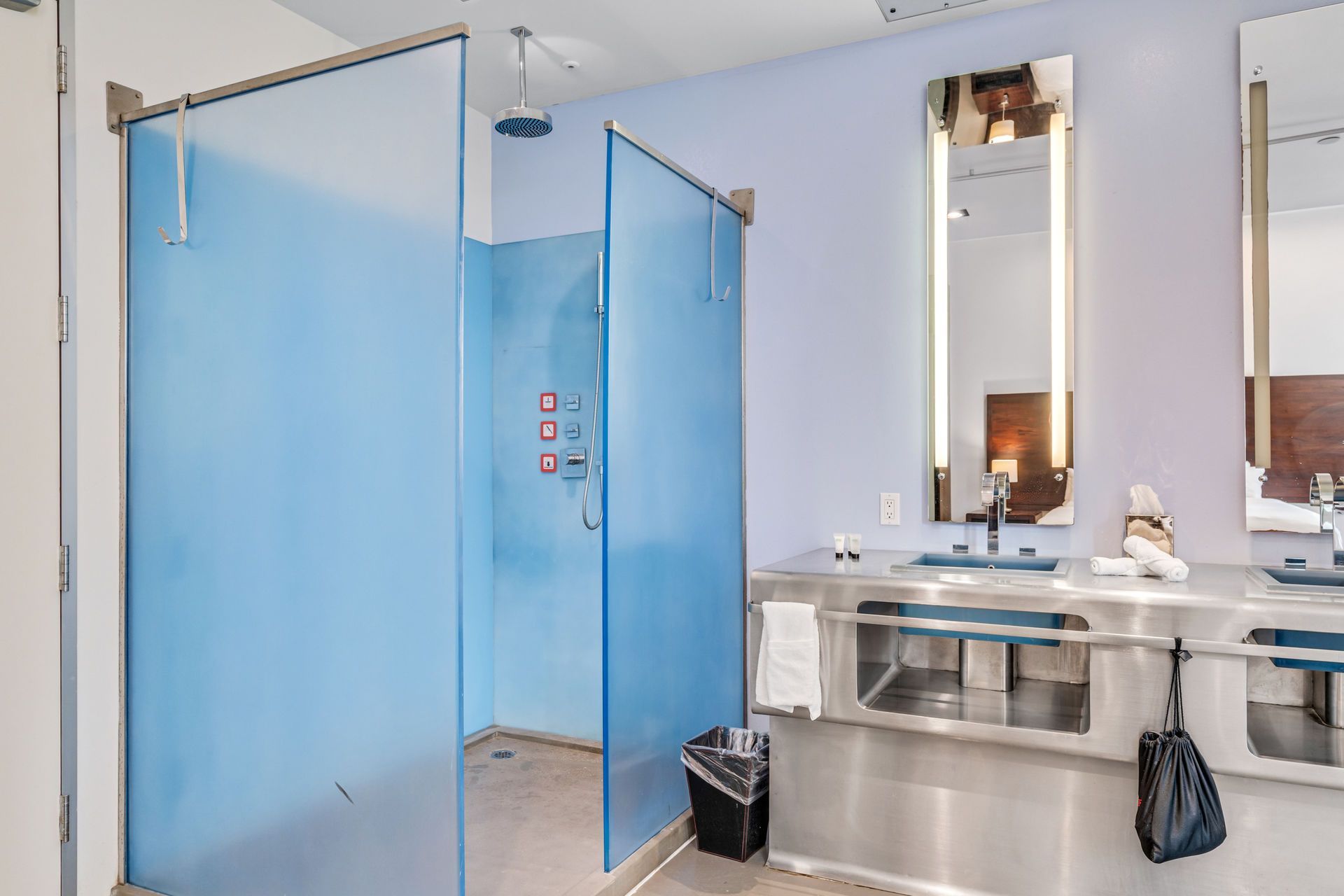 Public bathroom with blue glass shower stall, stainless steel sinks, mirrors, and waste bin.