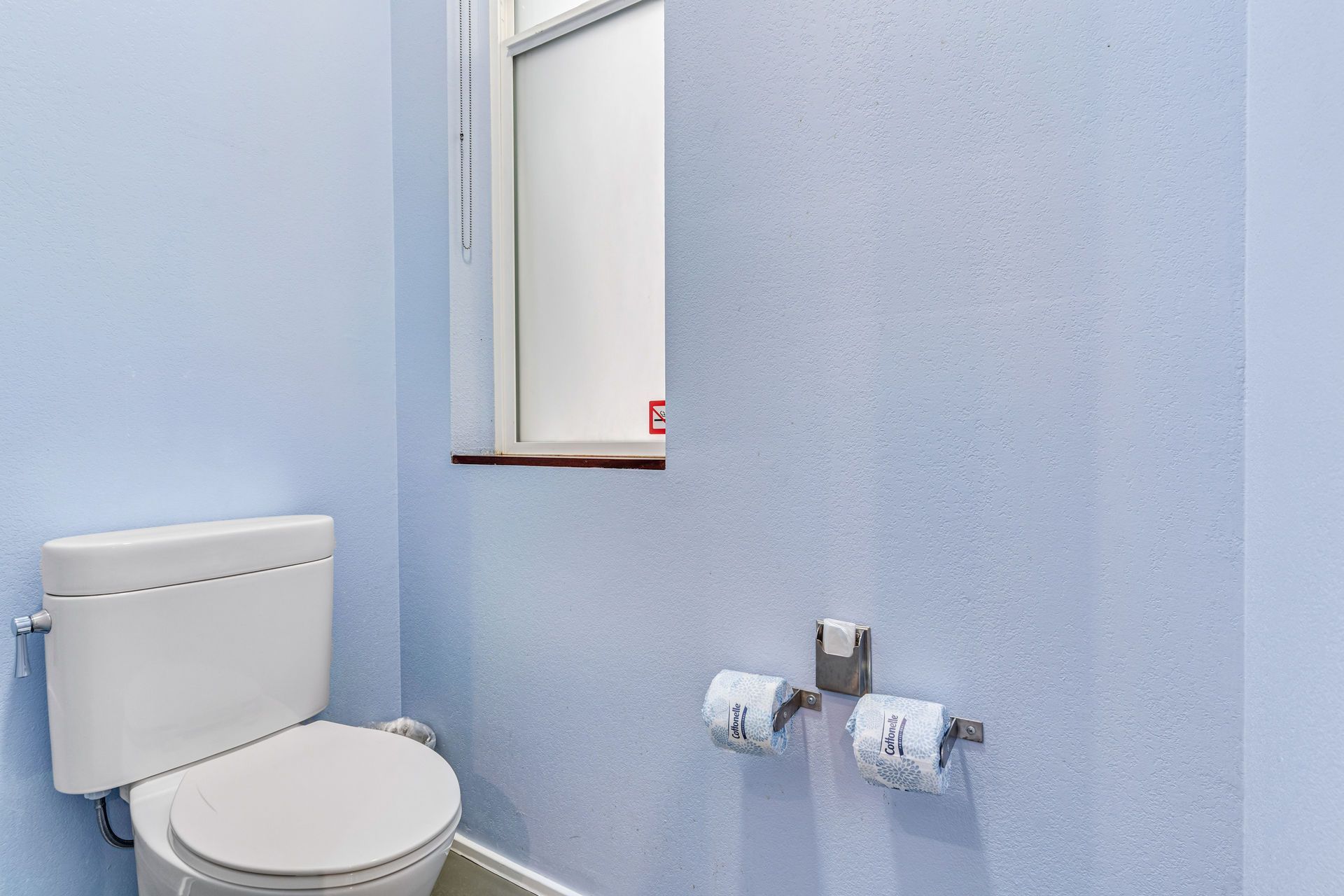 Blue bathroom with a toilet, window, and two rolls of toilet paper.