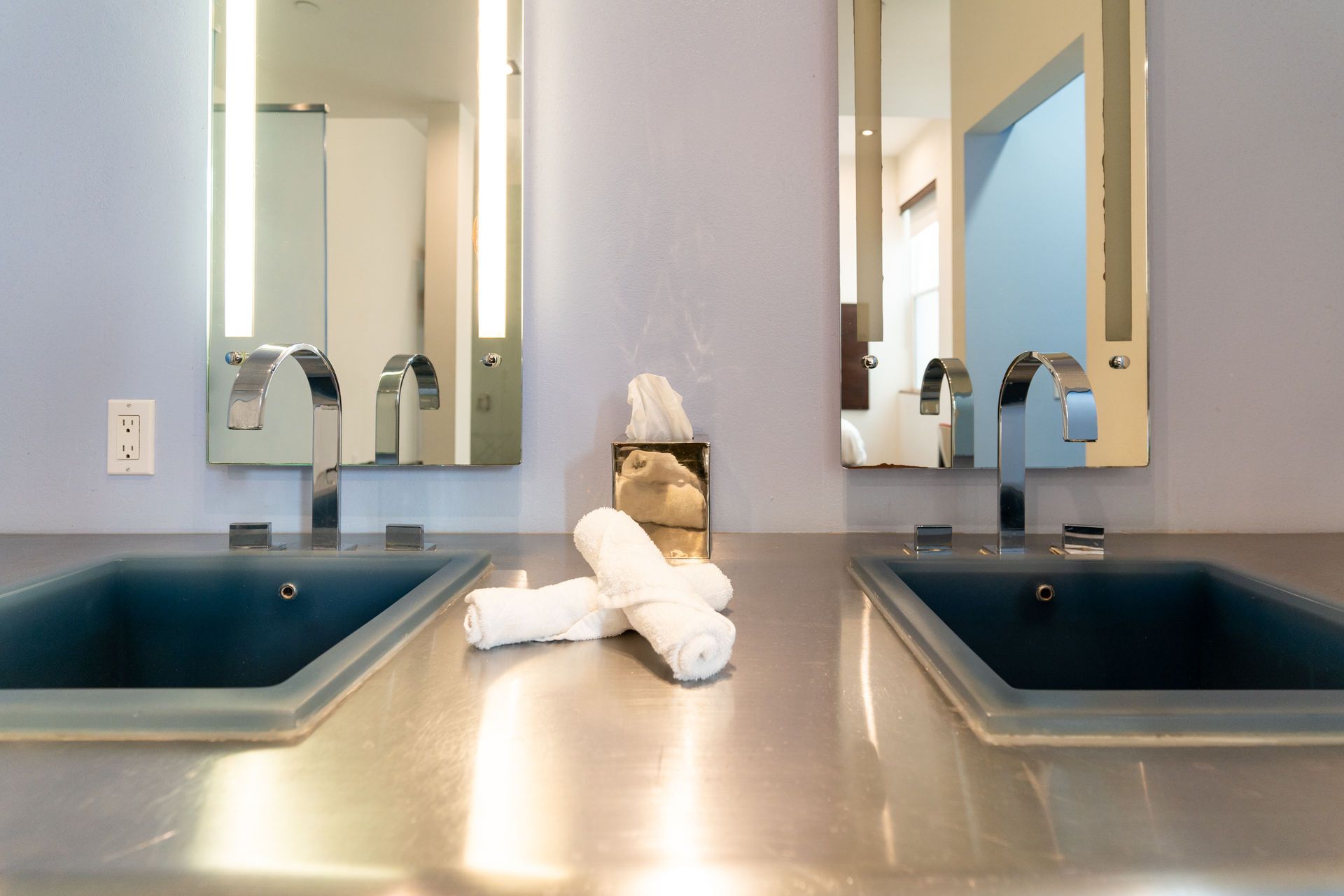 Two sinks with chrome faucets, mirrors with lights, and rolled white towels on a metal countertop.