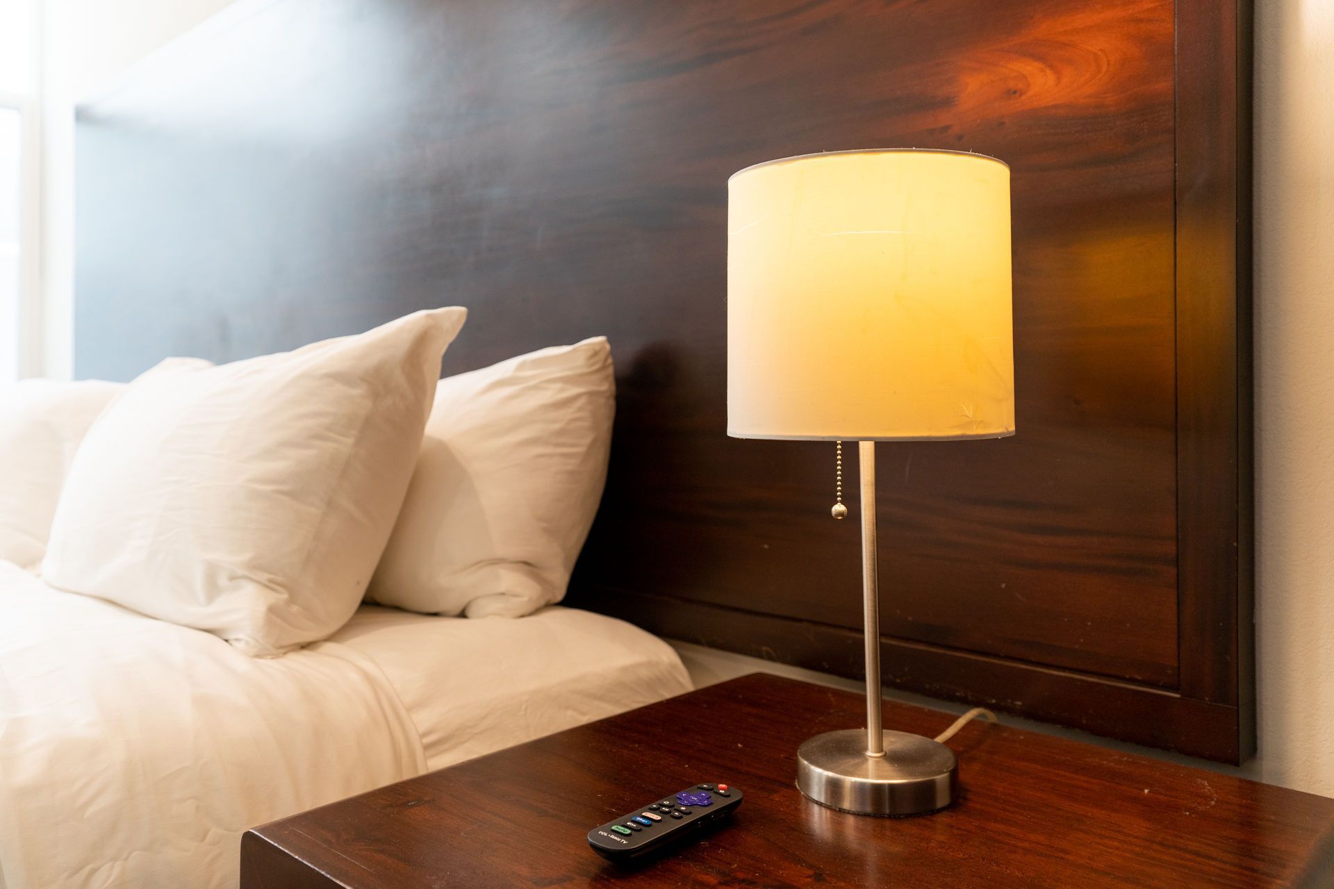 Bedside table with lamp and remote, beside a bed with white pillows and a dark headboard.
