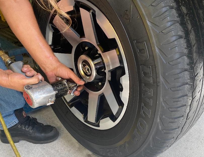 Person using an air wrench to remove a lug nut from a car tire, near a black rim.