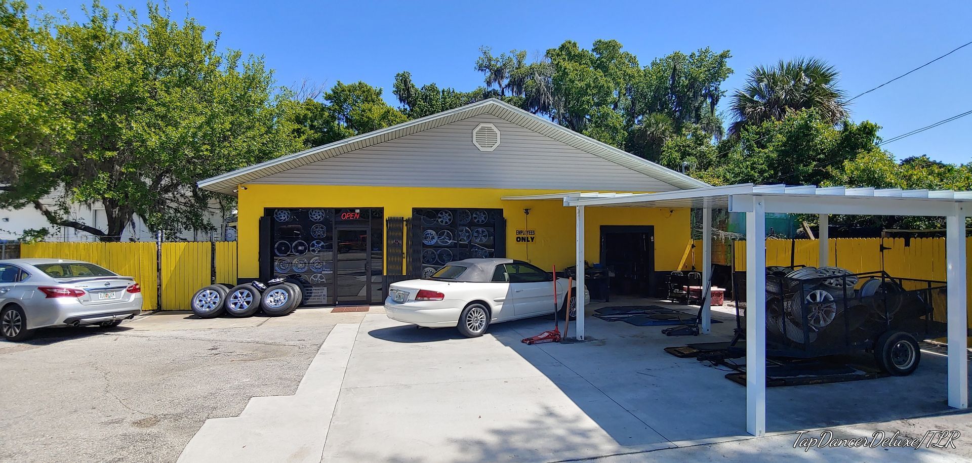 Yellow auto repair shop with cars and tires in driveway, trees and blue sky in background.