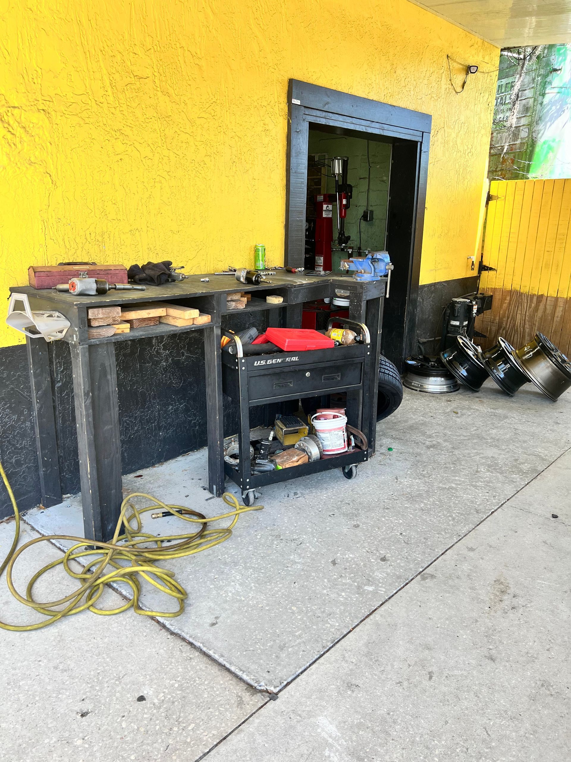 Black work cart with tools outside a shop; yellow wall, concrete floor, and wheels.