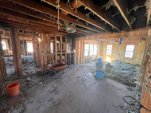 Damaged room with exposed beams, debris on floor, and wheelbarrow near bright windows