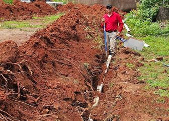 Worker in red shirt standing in a muddy trench beside piles of red soil, holding a bucket and tool.