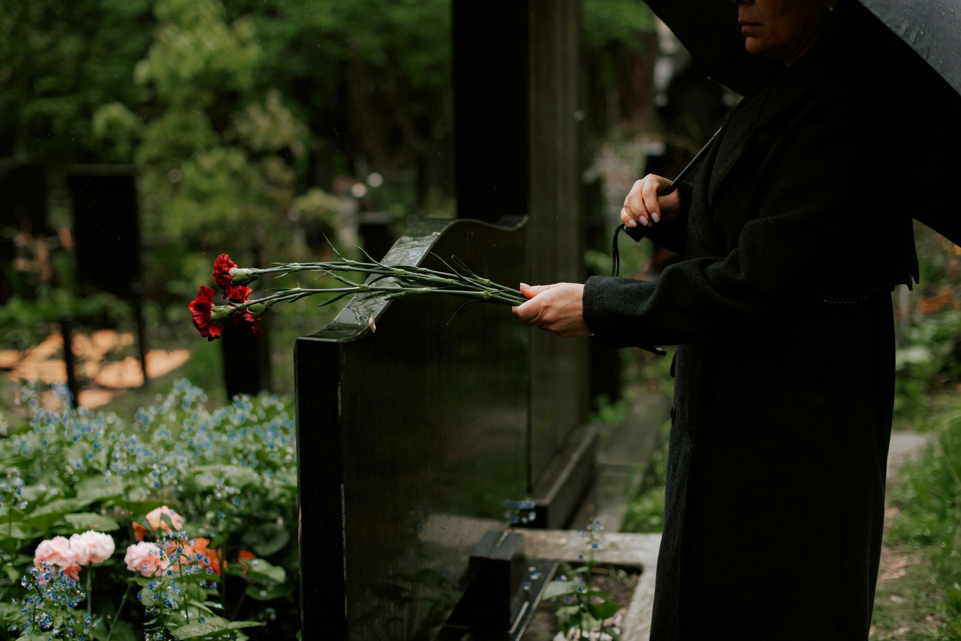 Person in black coat holds flowers, standing by a tombstone in a cemetery.