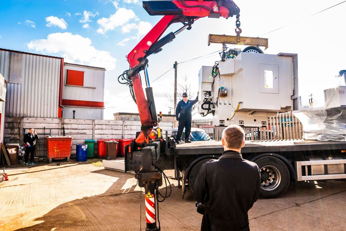 A crane lifts a large industrial machine onto a flatbed truck; a worker directs it.