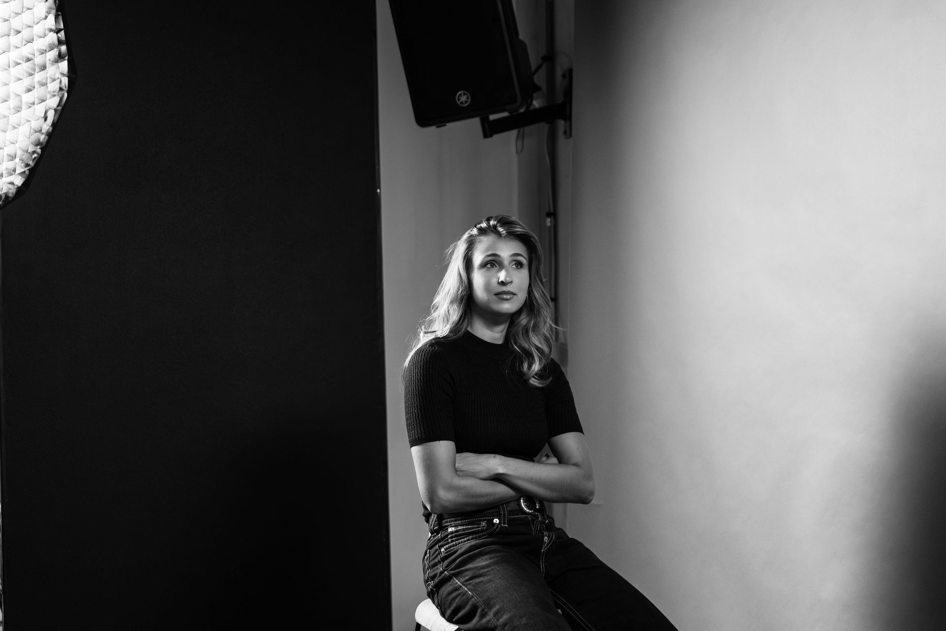 A person sits with arms crossed in a studio against a split black and white background, looking off to the side.