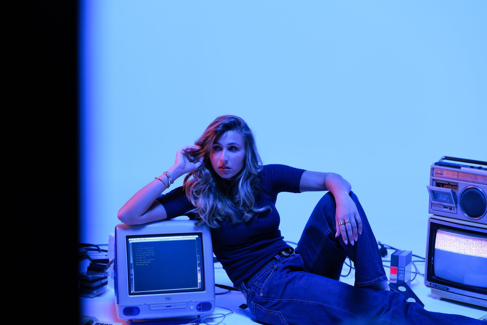A person sits on the floor in blue light, resting an arm on a vintage computer monitor next to a retro boombox.