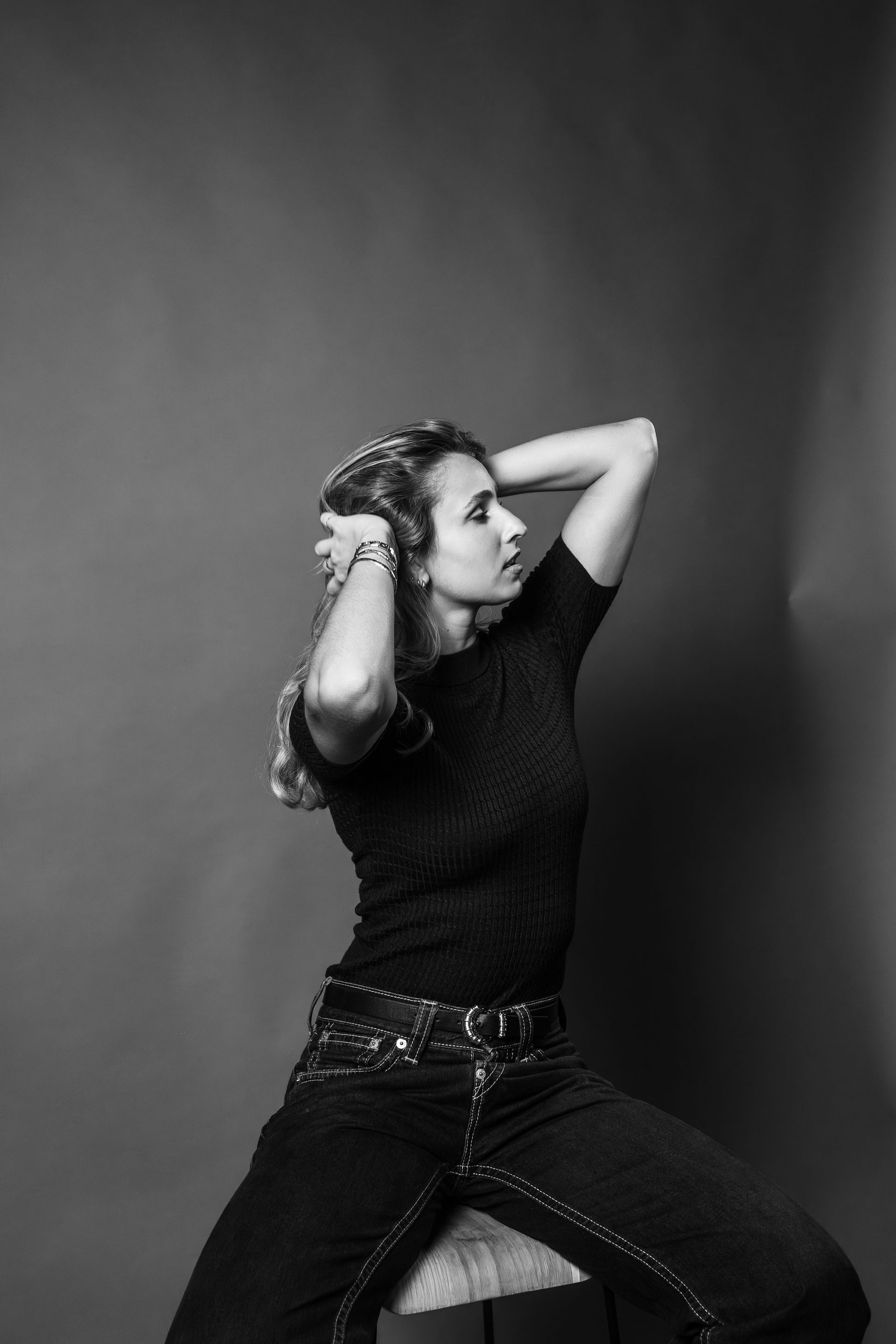 A person in a black t-shirt and jeans sits against a gray background, hands raised to their hair in a side profile view.