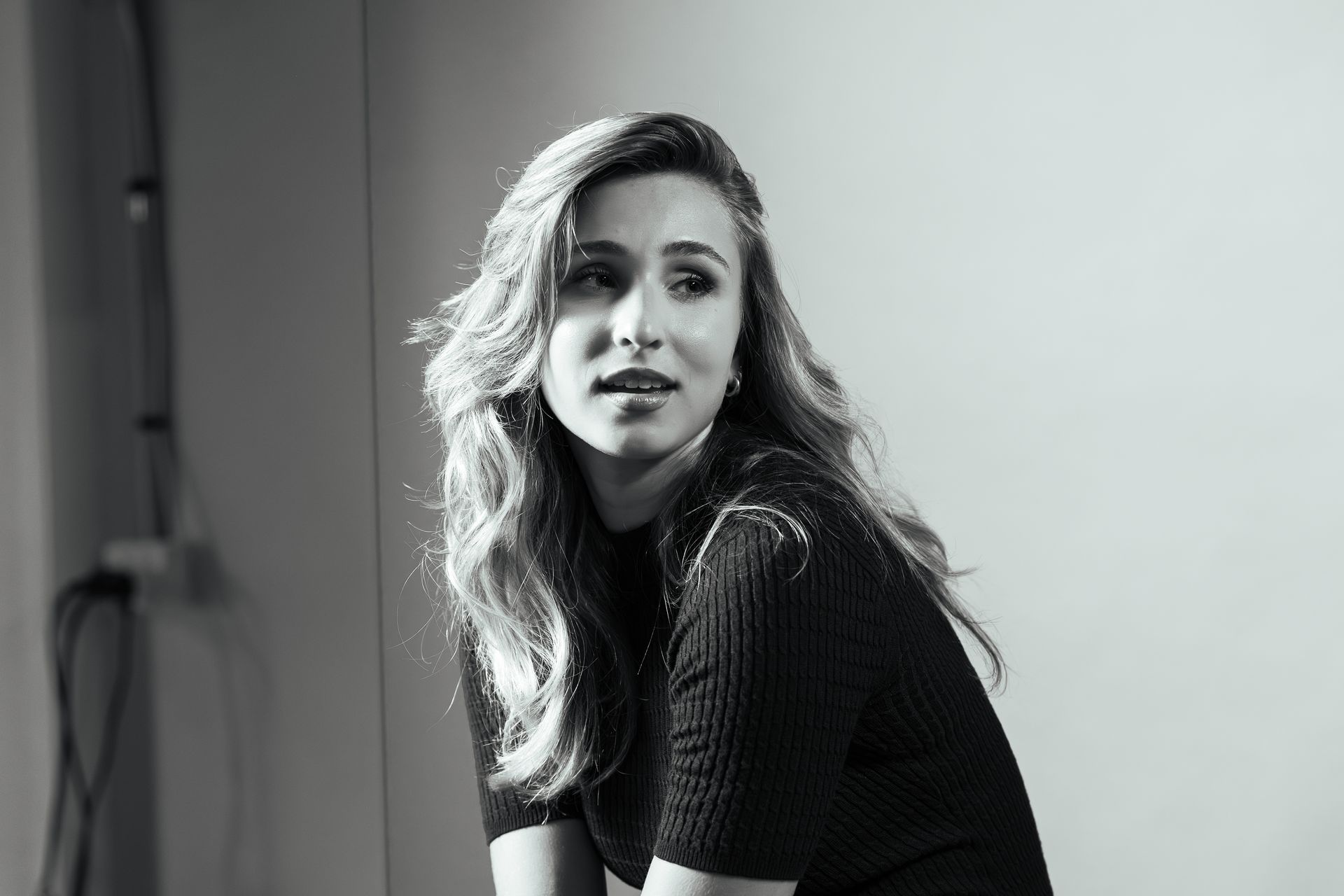 A black-and-white portrait of a person with long wavy hair looking to the side, wearing a ribbed top in a minimalist space.