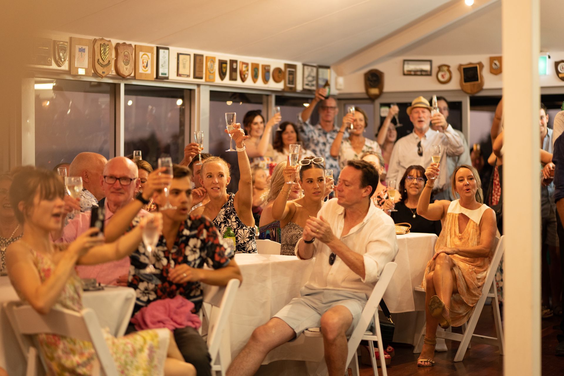 A large group of people are sitting at tables and chairs toasting with wine glasses.