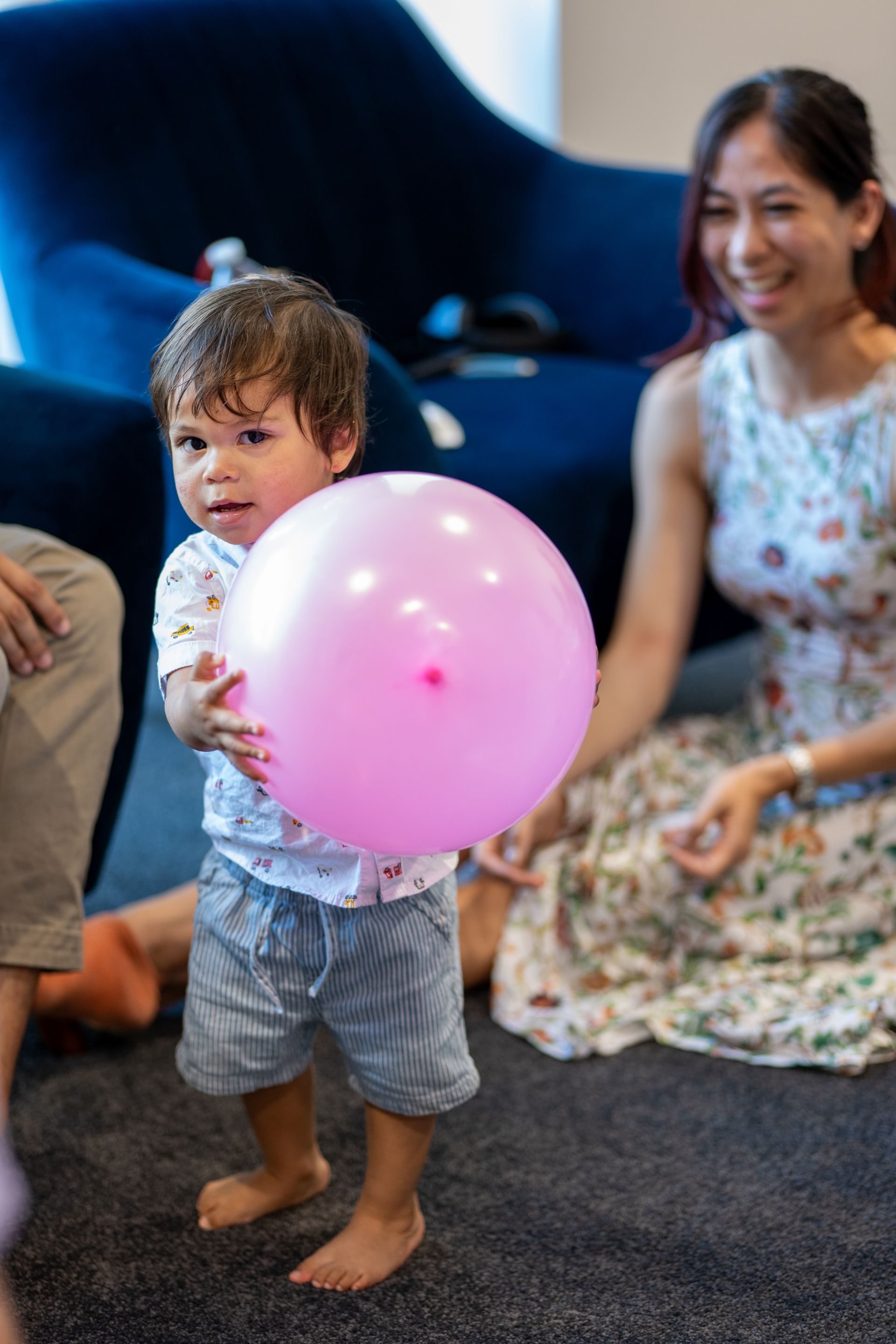 A little boy is holding a pink balloon while a woman sits on the floor.