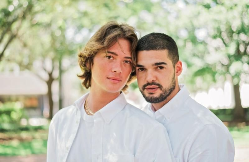 Two people in white shirts standing outdoors in a sunlit park, looking at the camera.