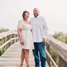 Two people standing on a wooden bridge, smiling in light-colored clothes