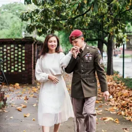 Woman in white dress and man in military uniform walking outdoors, holding hands amid fallen leaves.