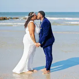 Couple in white dress and blue suit kissing on a sunny beach, holding hands at the shoreline