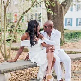 Couple in white outfits sharing a kiss on a stone bench in a leafy outdoor setting.