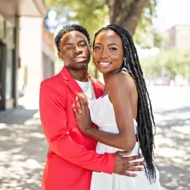 Couple embracing outdoors, smiling at the camera; one wears a red jacket and the other a white dress.