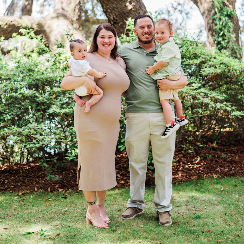 Family of four smiling outdoors in a garden, holding two toddlers, with trees and shrubs in the background