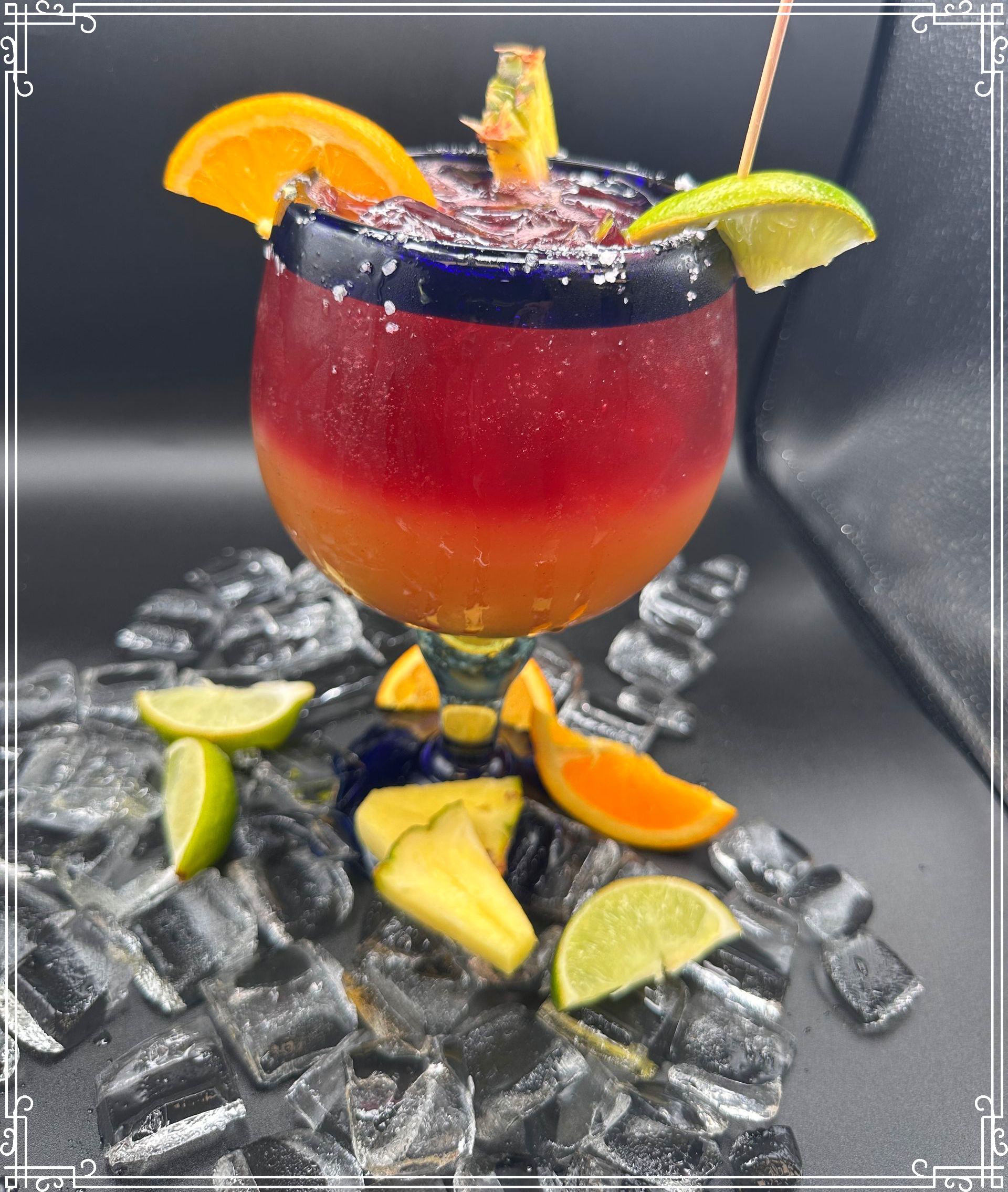 Cocktail in a blue-rimmed glass with orange and lime garnishes, surrounded by ice and fruit slices.