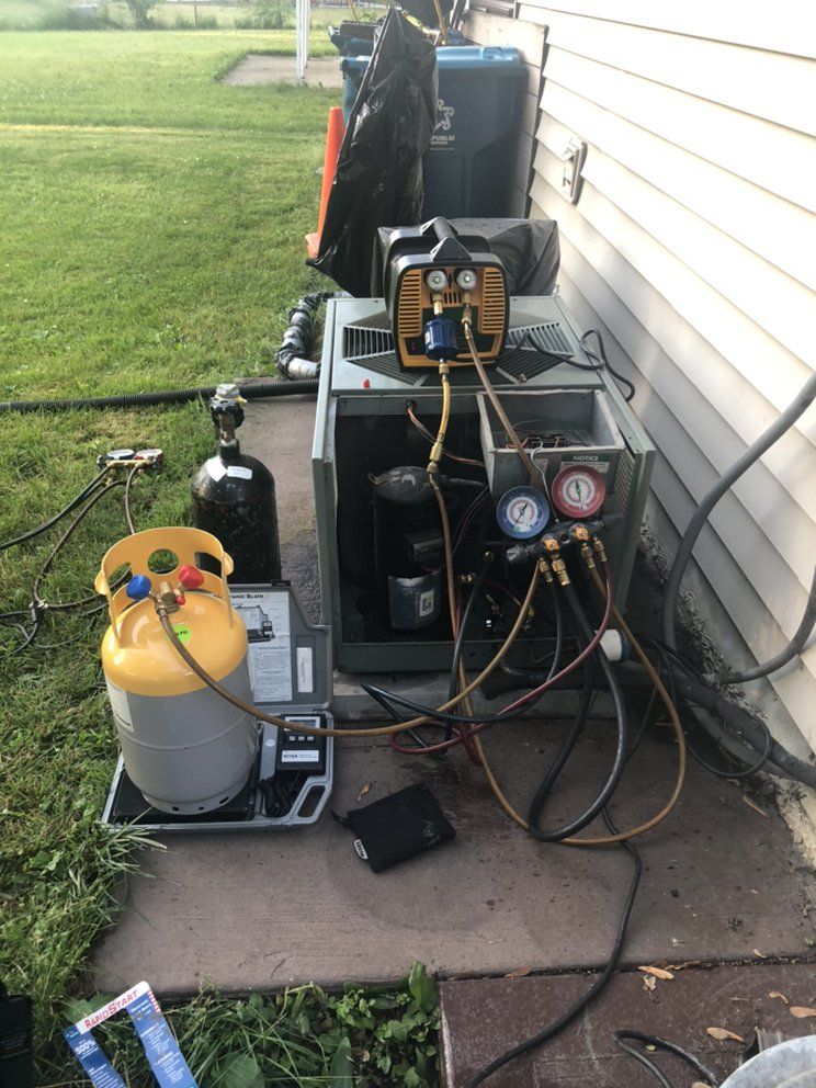 HVAC equipment outdoors; a technician working on a unit with gauges and tanks, near a house with green grass.