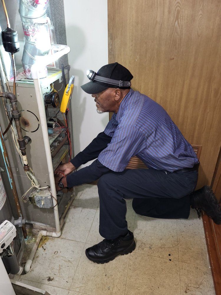 A technician kneels, examining a furnace, illuminated by a headlamp. He wears a blue shirt, black hat and shoes.