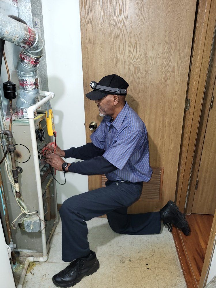 HVAC technician kneels, inspecting a furnace. He wears a hat with a light and a blue striped shirt.