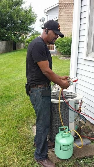 HVAC technician works on an AC unit outdoors. He connects hoses to a green refrigerant tank.