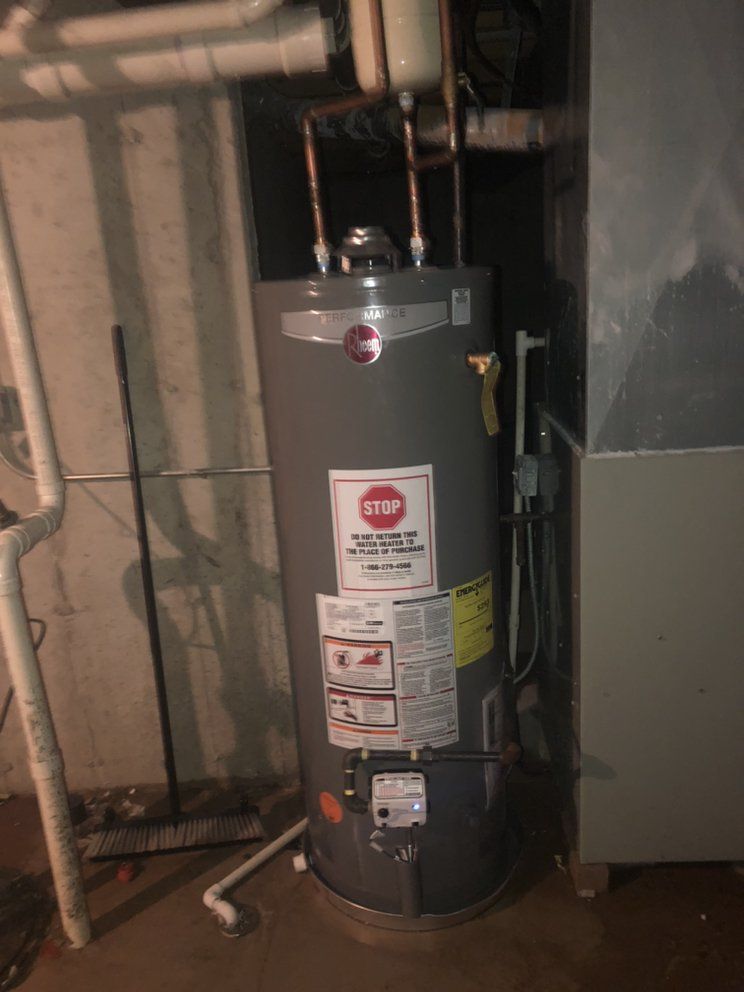 A vertical Rheem water heater in a basement, next to a furnace. Gray tank, with pipes and warning labels.