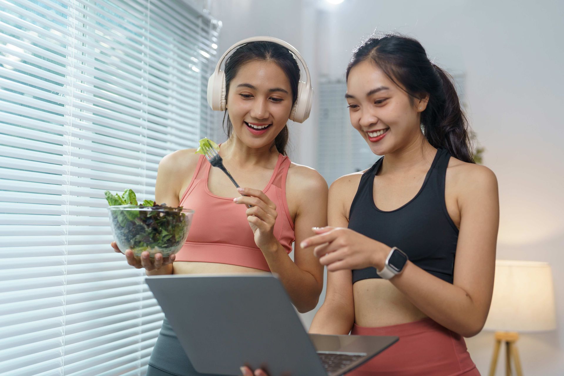 Two women in workout clothes, one holding salad, looking at a laptop with happy expressions.