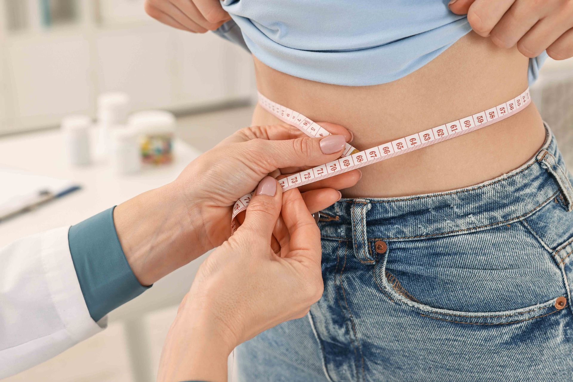 Doctor measuring a person's waist with a measuring tape. The person wears jeans and a blue shirt, in a medical office.