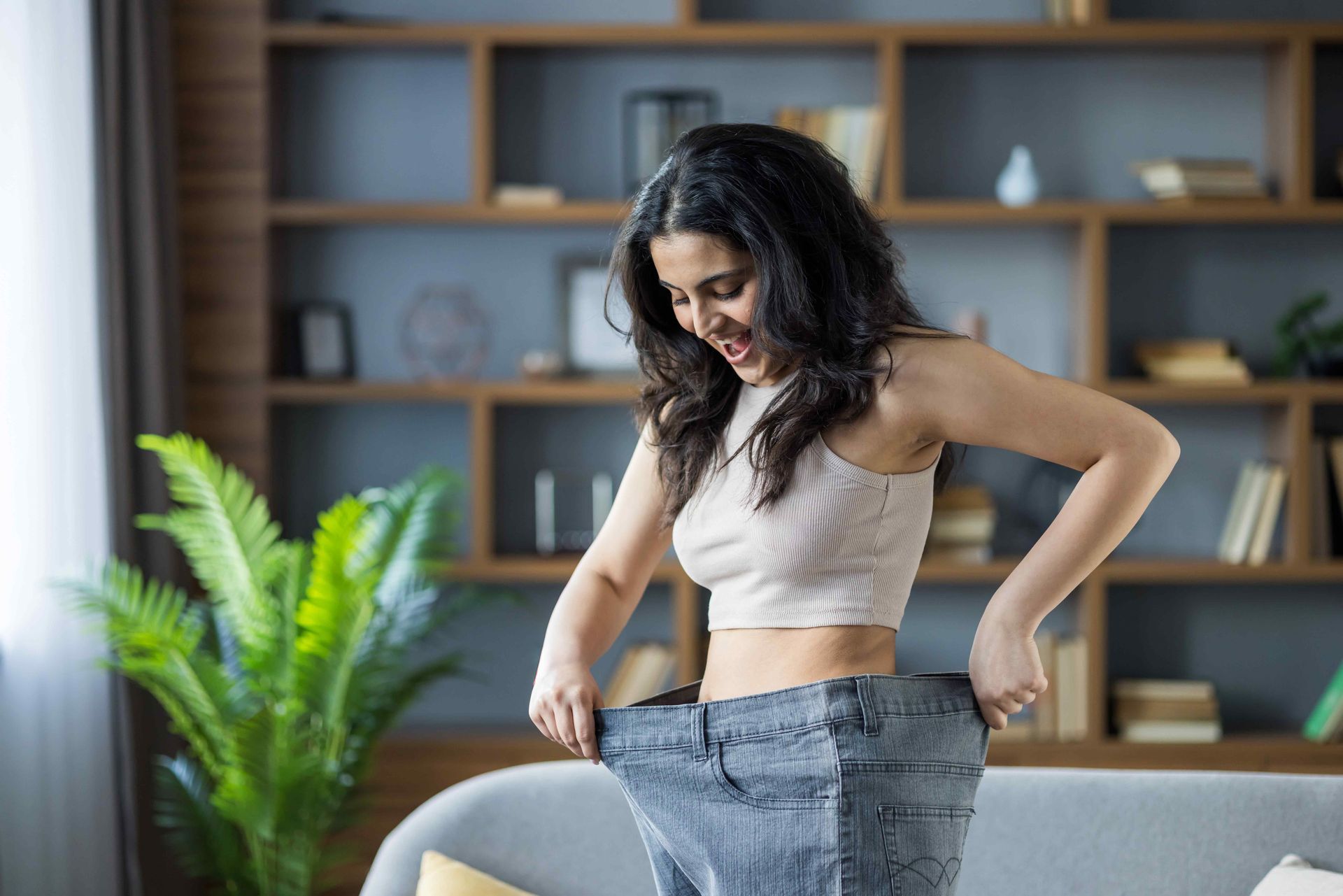 Woman smiling, wearing oversized jeans, showing weight loss in living room.