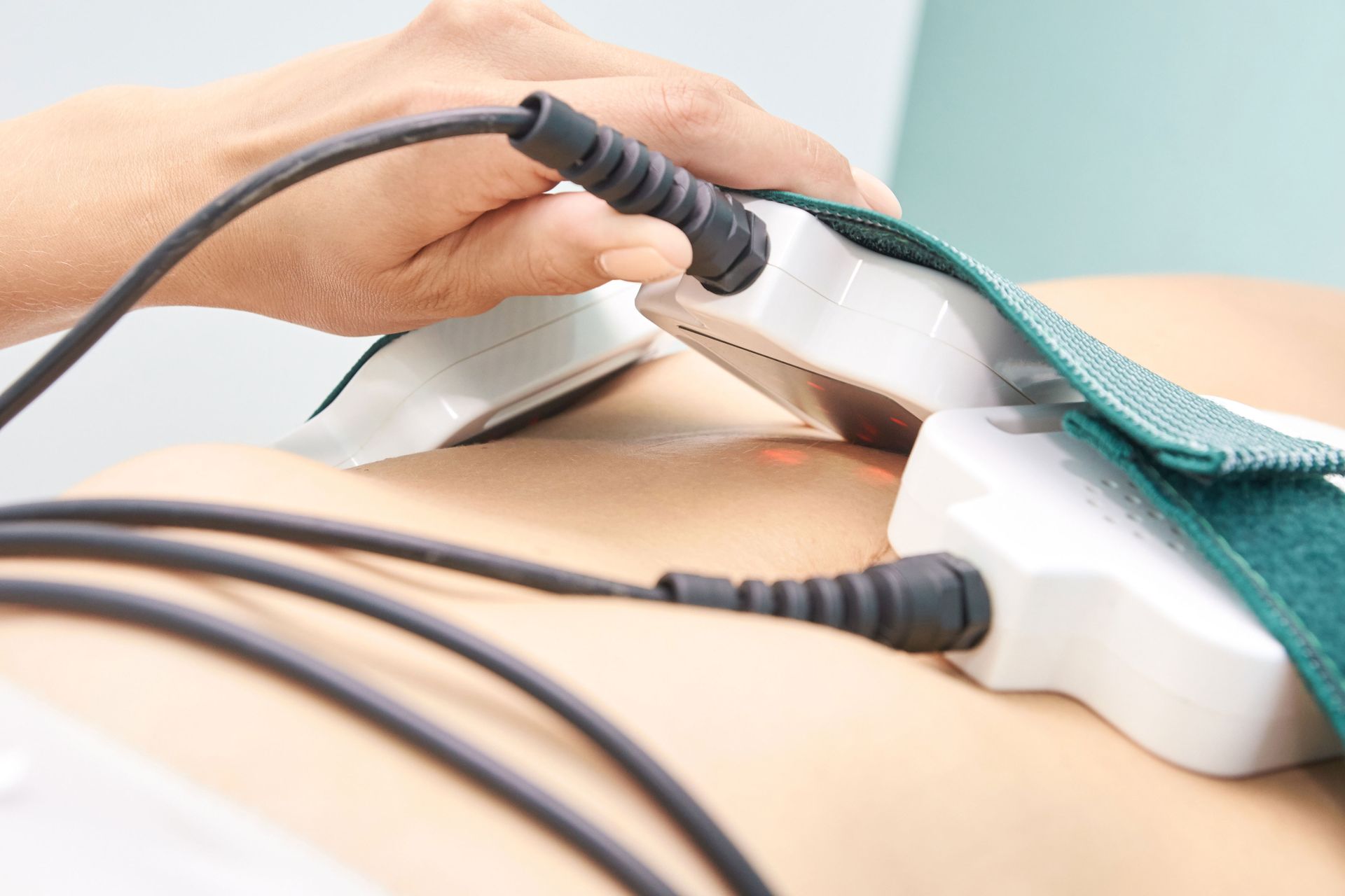 Person's abdomen with electrotherapy pads attached, held in place by green straps. Cables connect to the pads.