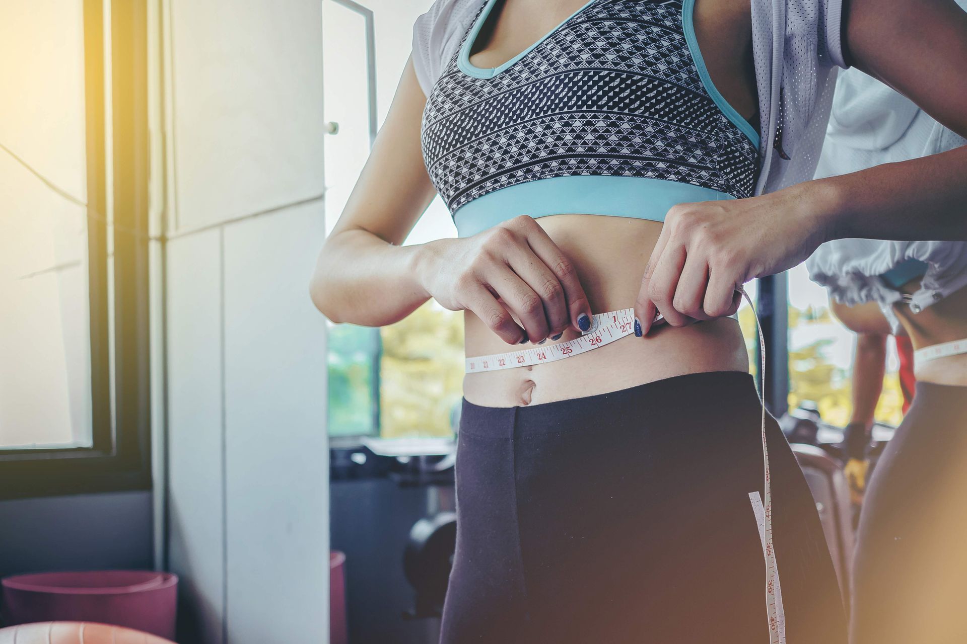 Woman measuring her waist with a white tape measure in a gym setting.