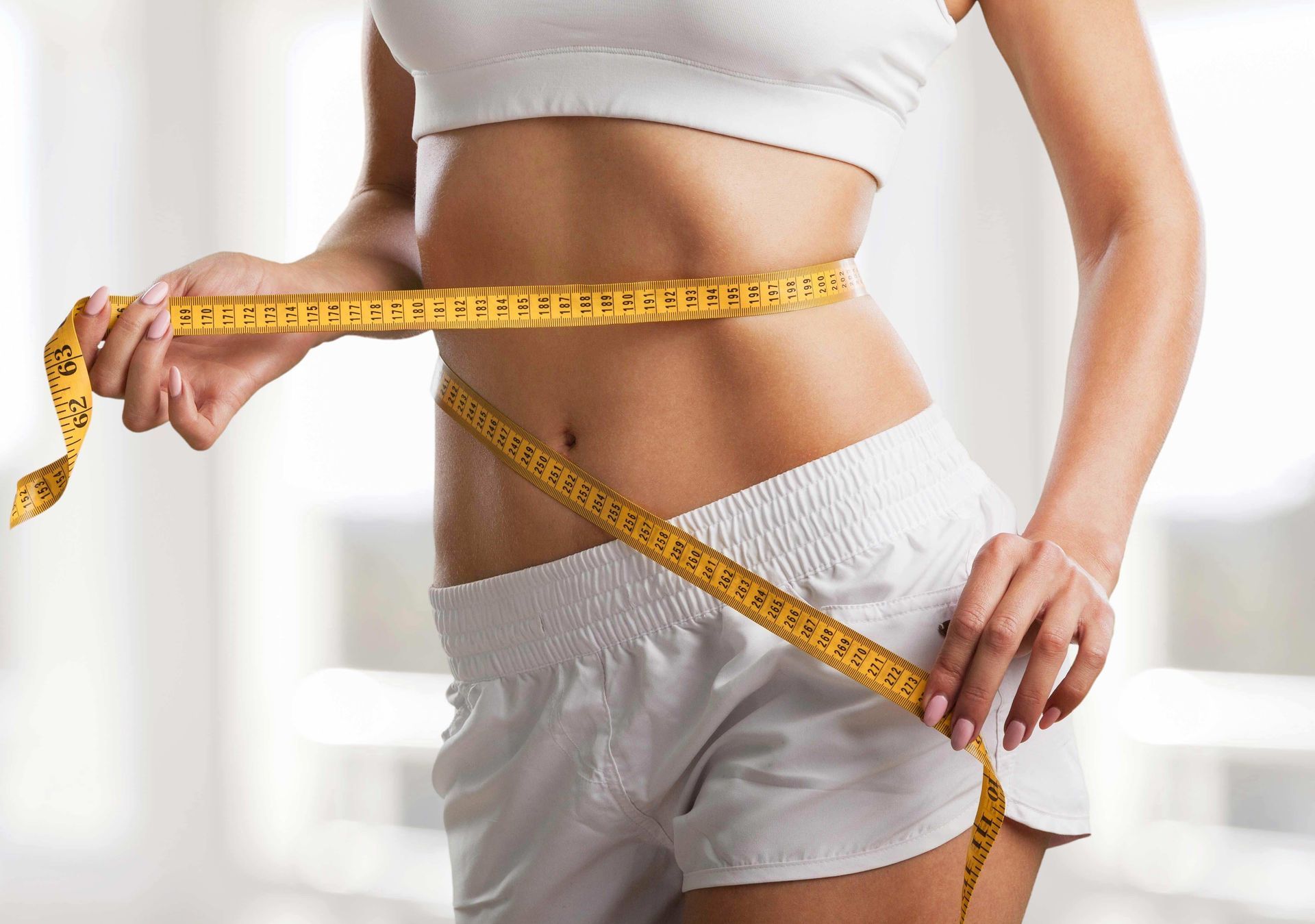 Woman in white shorts and top measuring waist with yellow tape measure.