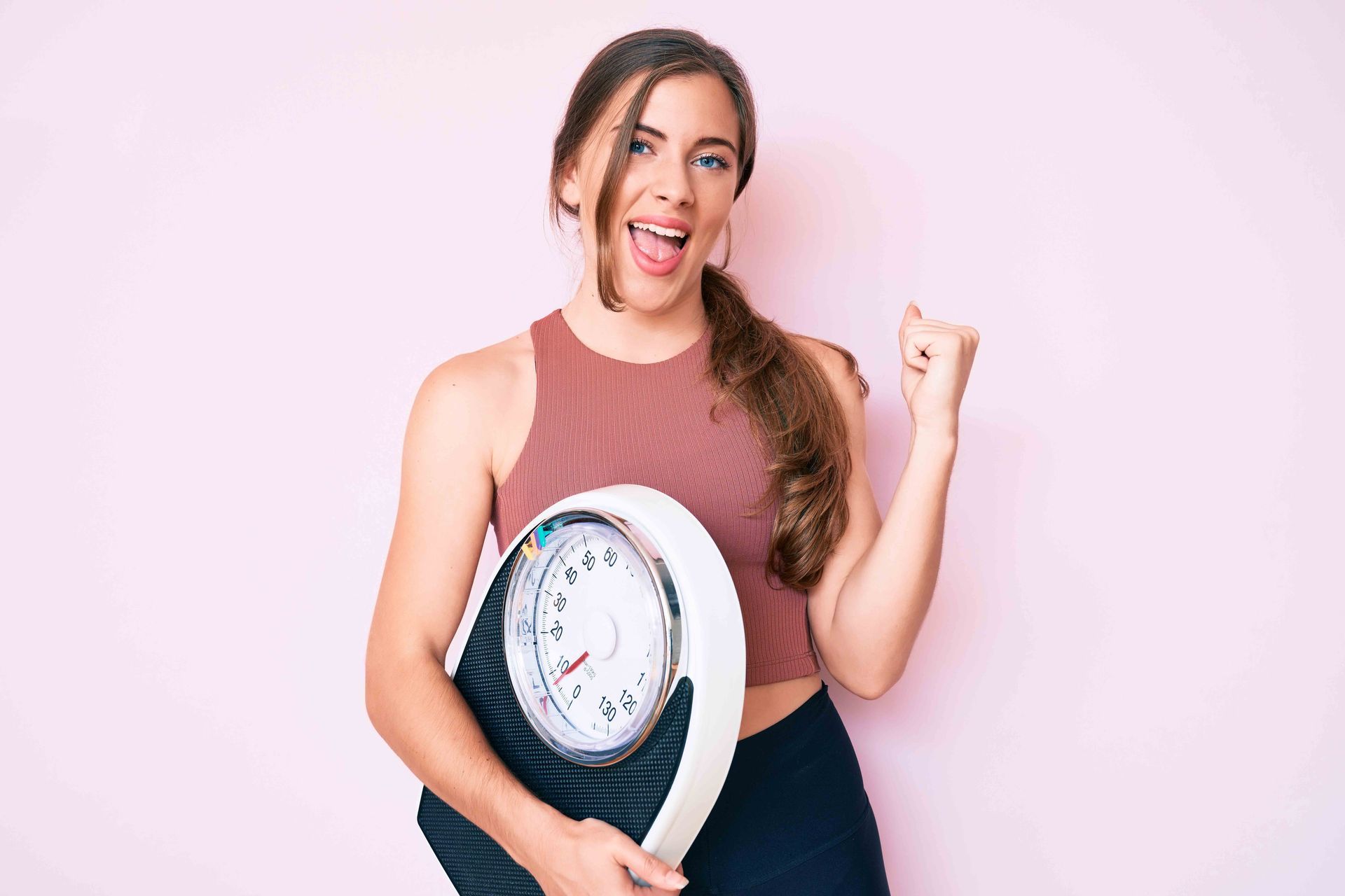 Woman holding a scale, smiling, and celebrating with her fist in the air, against a pink background.