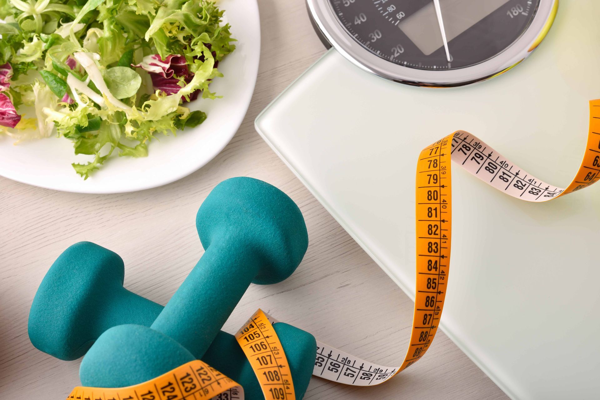 Salad, dumbbells, tape measure, and scale representing healthy eating and exercise.