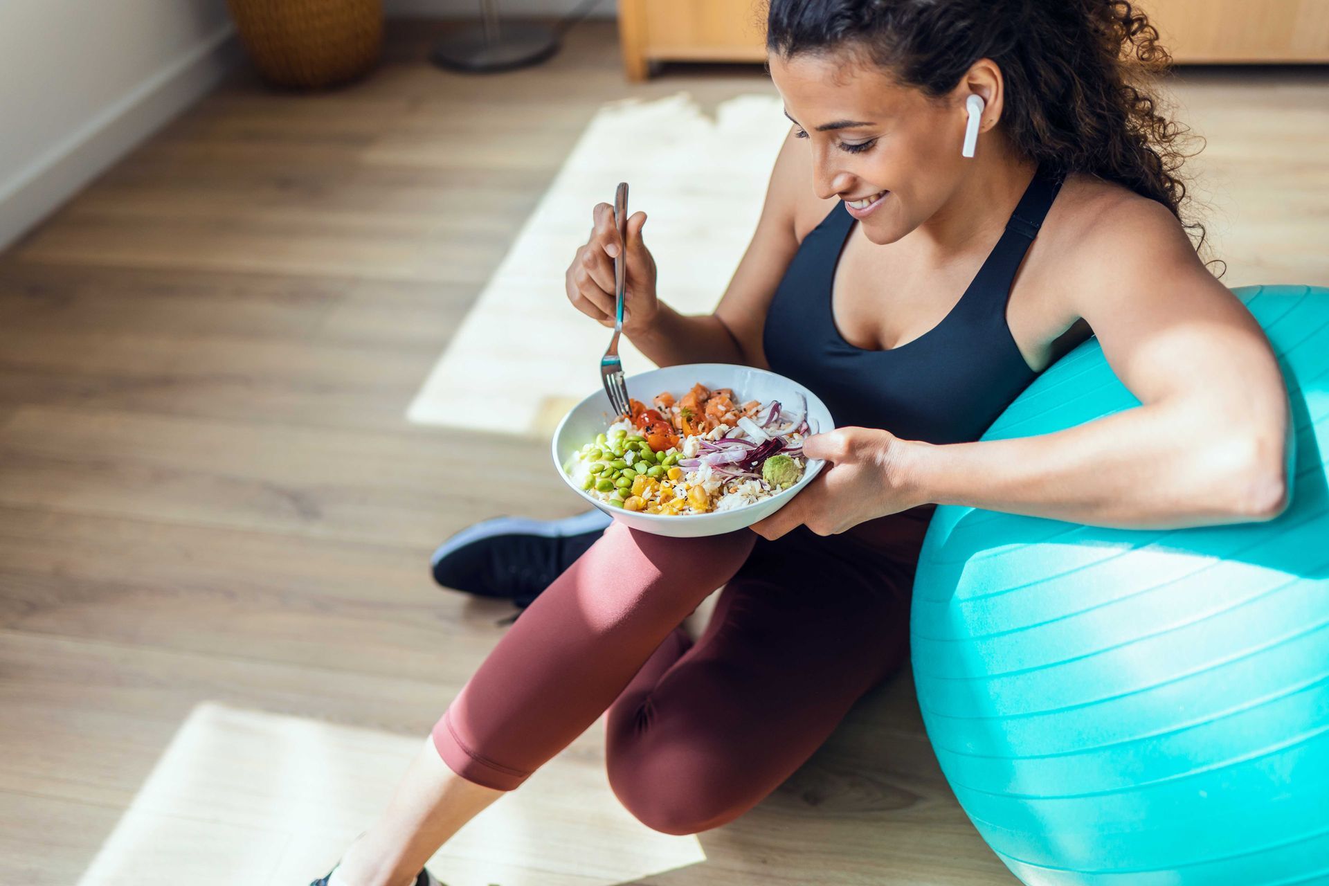 Woman in athletic wear eats a meal while leaning on a blue exercise ball.