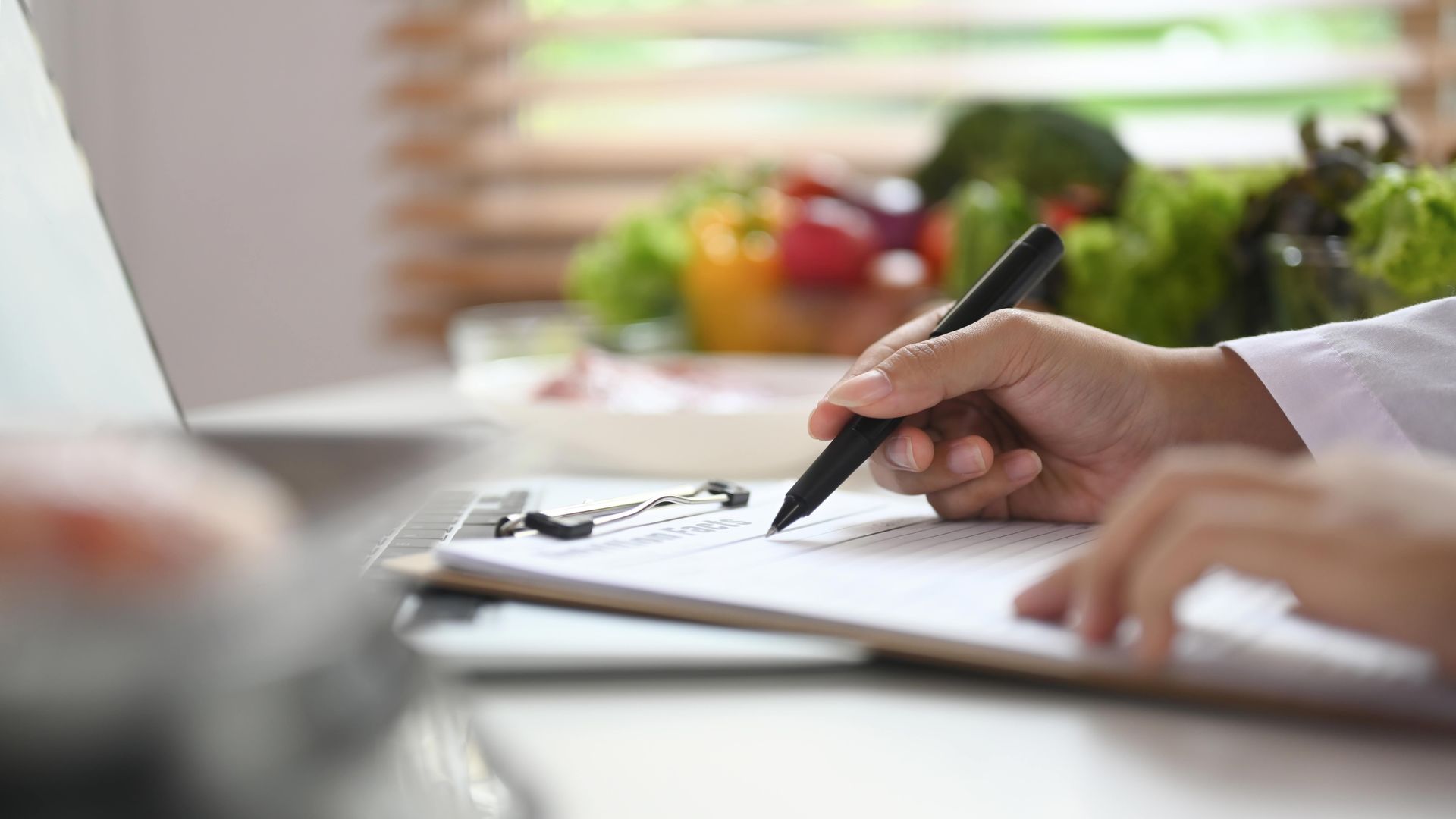 Hands writing on a clipboard with a pen, vegetables in background near a laptop.