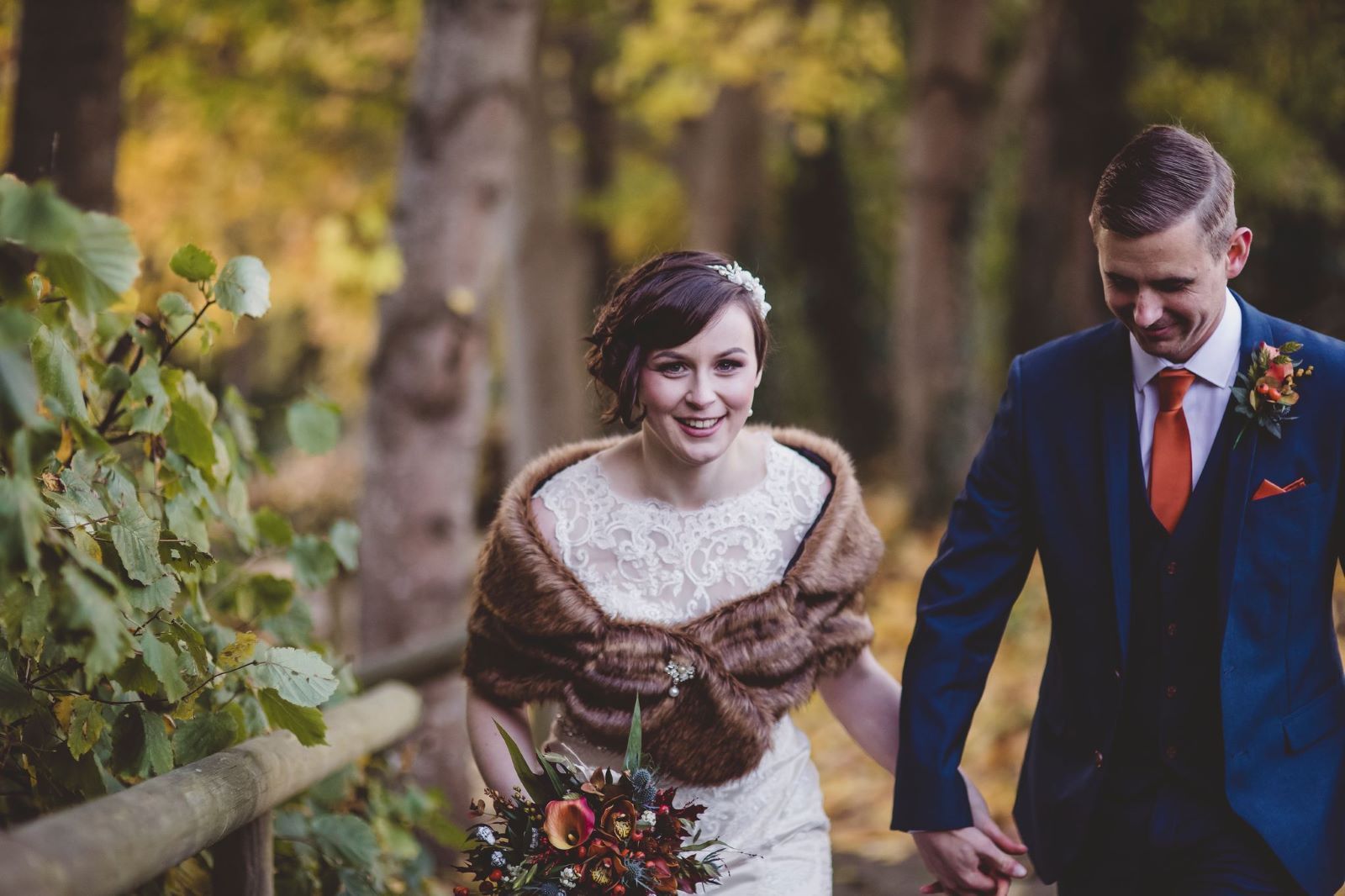 A bride and groom are walking through the woods holding hands.