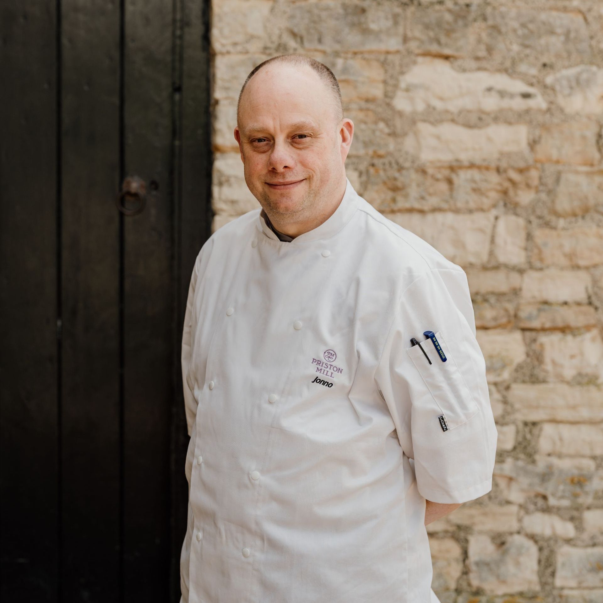 A man in a chef 's jacket is standing in front of a stone wall.