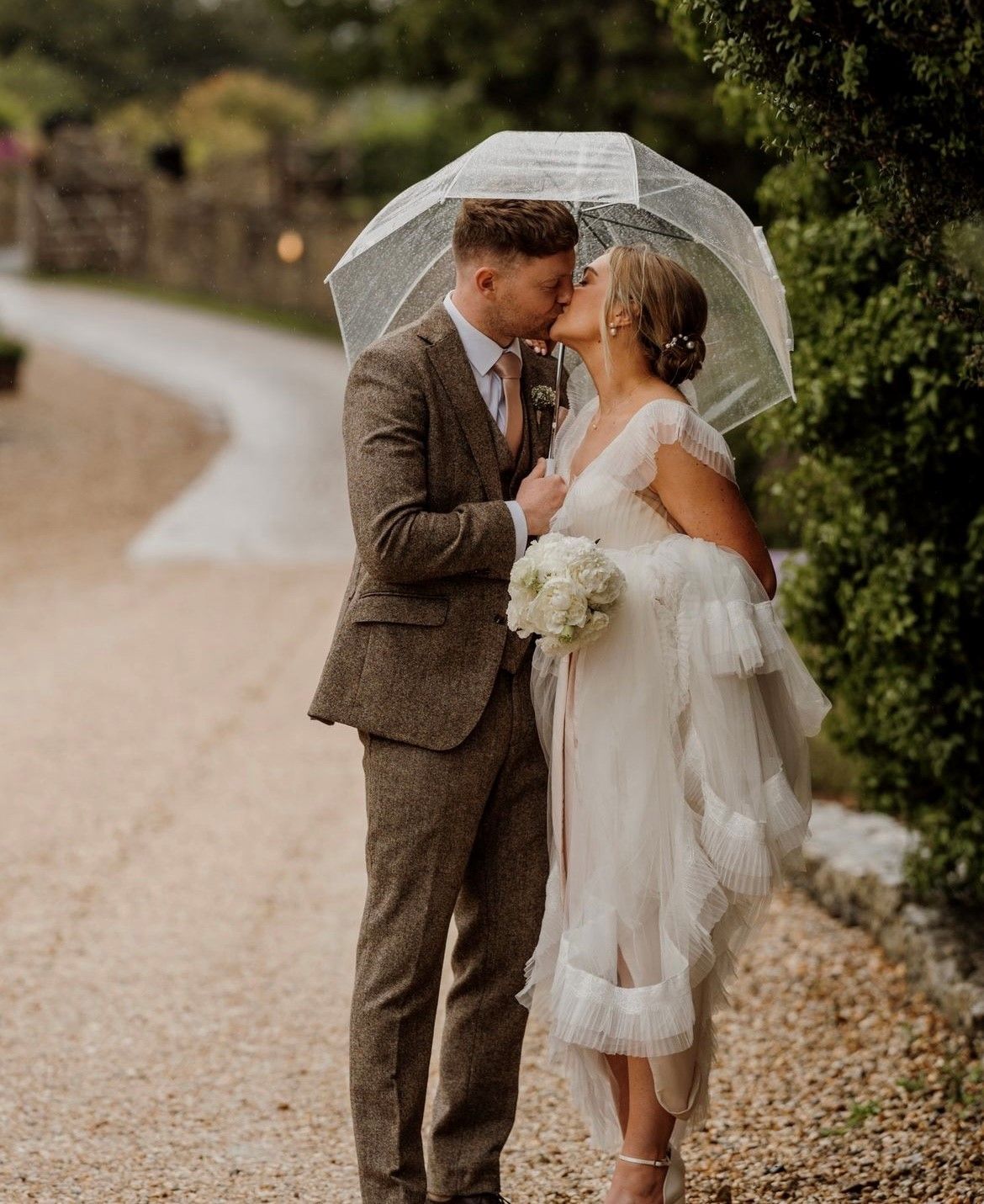 A bride and groom kissing under an umbrella on a gravel road.