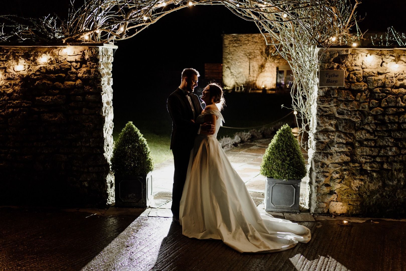 A bride and groom are standing under an archway at night.