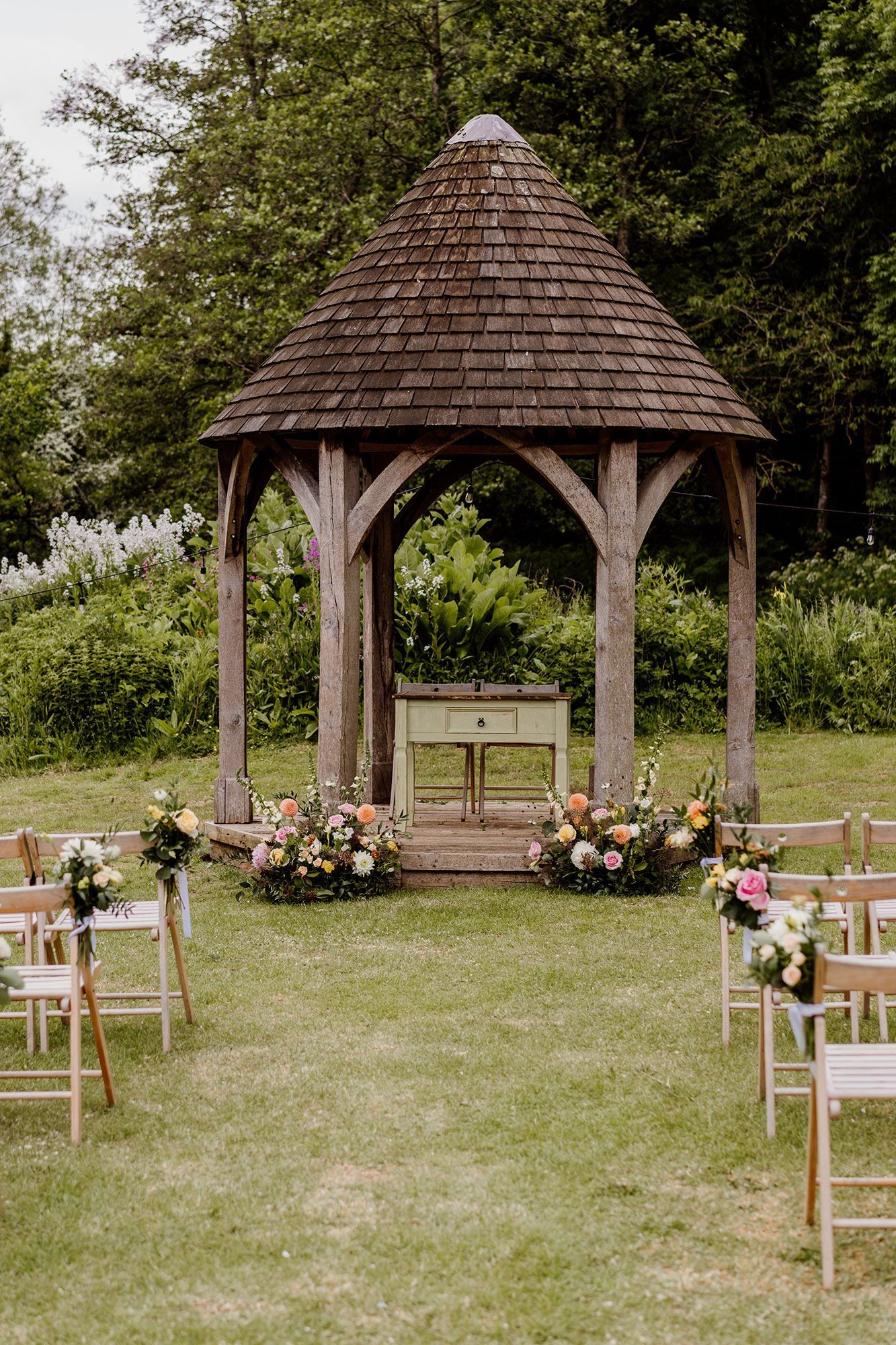 A wooden gazebo is decorated with flowers and chairs for a wedding ceremony.