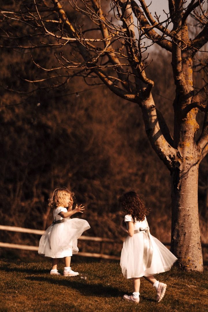 Two little girls in white dresses are standing next to a tree.